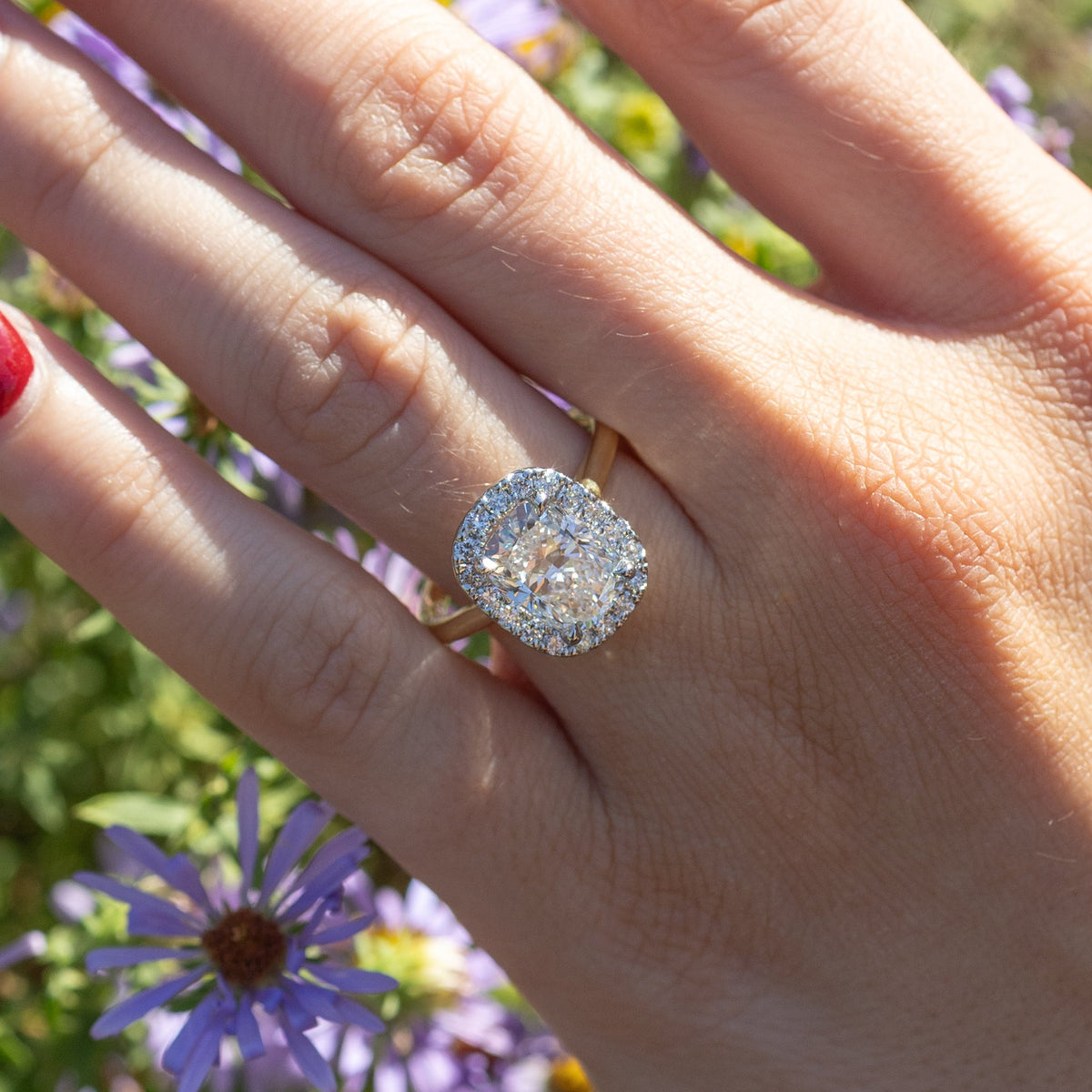 A close up shot of a large elongated cushion diamond engagement ring. The center diamond is surrounded by a halo of small prong-set round diamonds. In the background is a hand and blurry purple and green flowers.