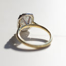 The back profile of a dual-tone elongated cushion diamond engagement ring. The band is yellow gold and the basket is platinum. It's sitting on a white marble backdrop with sharp shadows.