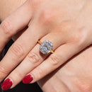 An elongated cushion engagement ring resting on the left ring finger of a hand with red nail polish. The ring is in direct sunlight and has a row of round diamonds in a halo formed around the center stone.