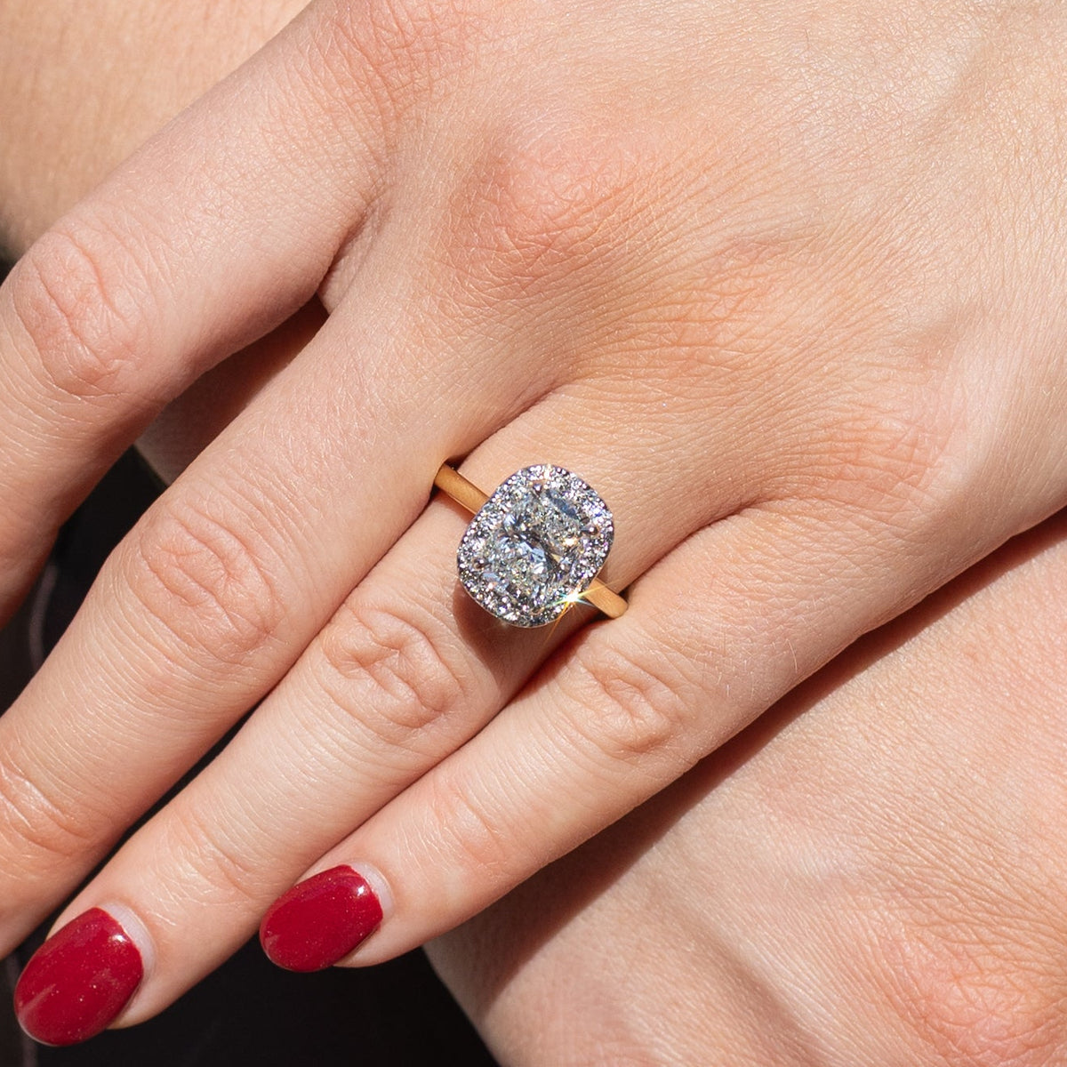 An elongated cushion engagement ring resting on the left ring finger of a hand with red nail polish. The ring is in direct sunlight and has a row of round diamonds in a halo formed around the center stone.