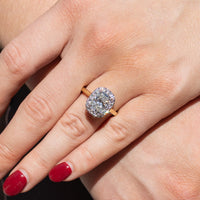 An elongated cushion engagement ring resting on the left ring finger of a hand with red nail polish. The ring is in direct sunlight and has a row of round diamonds in a halo formed around the center stone.