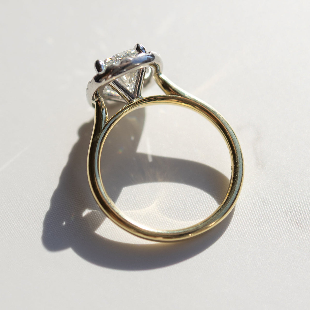 The side profile view of a dual-tone engagement ring with a yellow gold band and platinum basket. The ring is on a white marble backdrop.