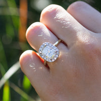 A close up shot of a dual-tone elongated cushion engagement ring on a ring finger. The cushion is surrounded by a halo of small round diamonds. In the background is blurry green grass.