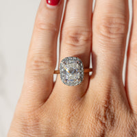 An on-hand shot of an elongated cushion diamond engagement ring with a halo. In the background is a blurry white backdrop.