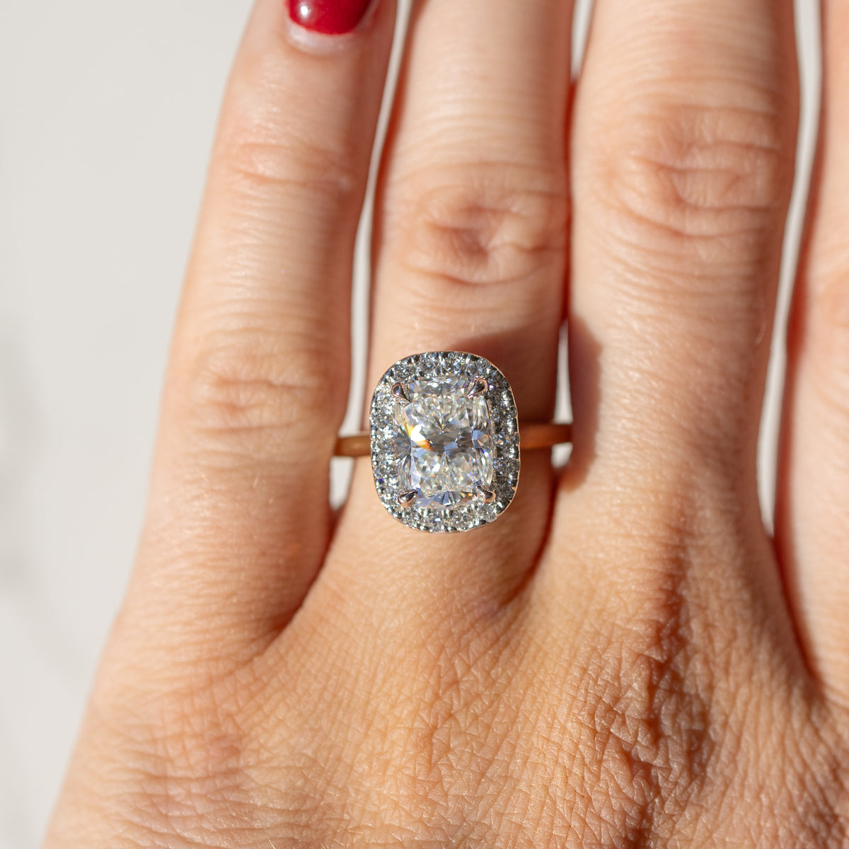 An on-hand shot of an elongated cushion diamond engagement ring with a halo. In the background is a blurry white backdrop.