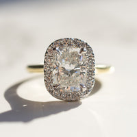 A large elongated cushion diamond engagement ring surrounded by a halo of round diamonds sitting on a white marble backdrop. The ring is yellow gold and platinum.
