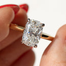 A close-up of a hand holding an elongated cushion cut diamond solitaire ring on a gold band against a clean white background.