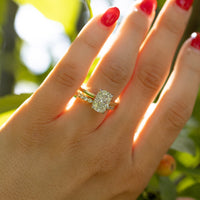 A close-up of a hand with red nail polish wearing a yellow gold ring with a large elongated cushion-cut diamond and a smaller diamond band, set against a blurred background of green leaves and sunlight.