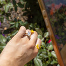 A close-up lifestyle shot of a hand wearing a dual-toned ring with a clear elongated cushion diamond, holding a smartphone to take a photo of yellow flowers in a garden with green foliage.