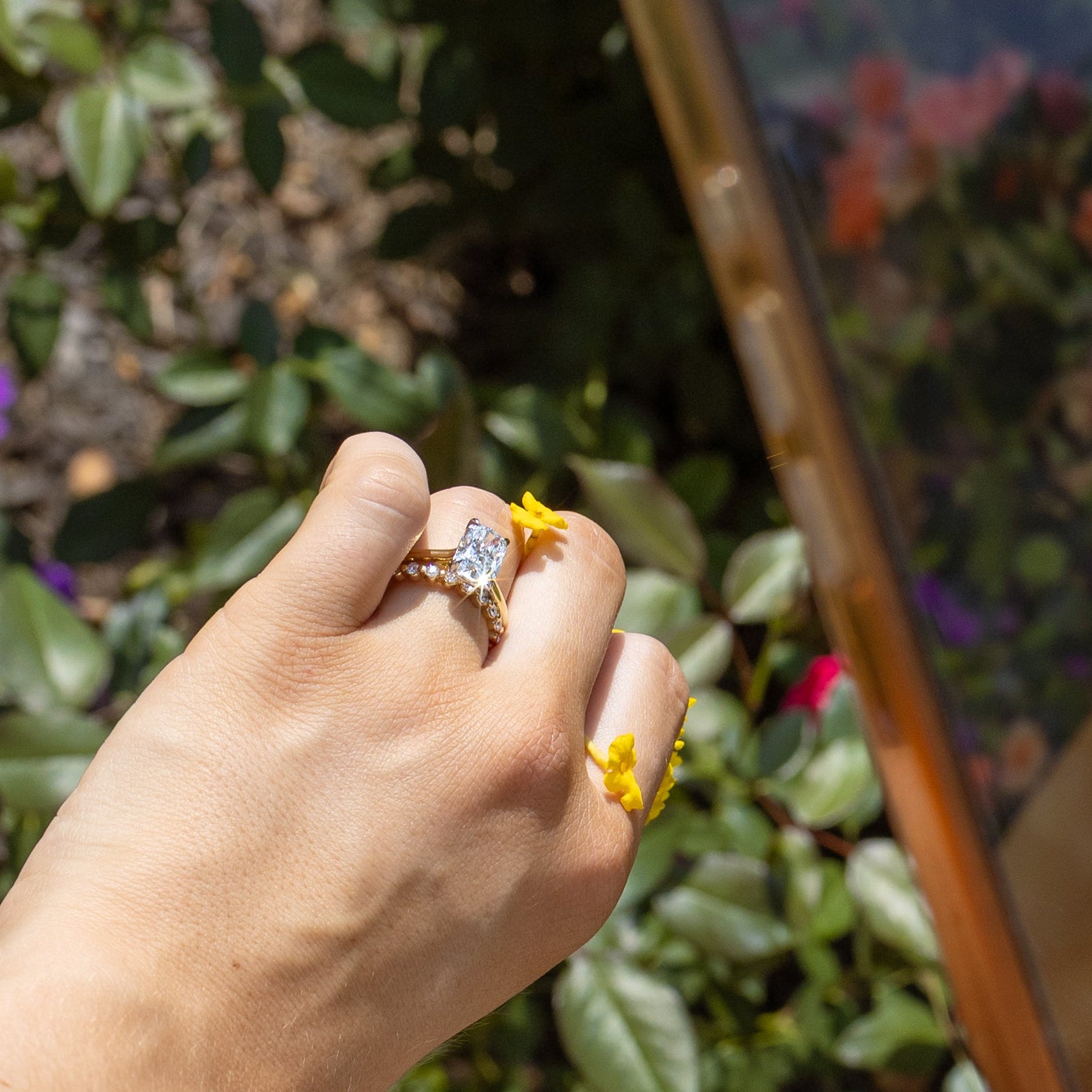 A close-up lifestyle shot of a hand wearing a dual-toned ring with a clear elongated cushion diamond, holding a smartphone to take a photo of yellow flowers in a garden with green foliage.