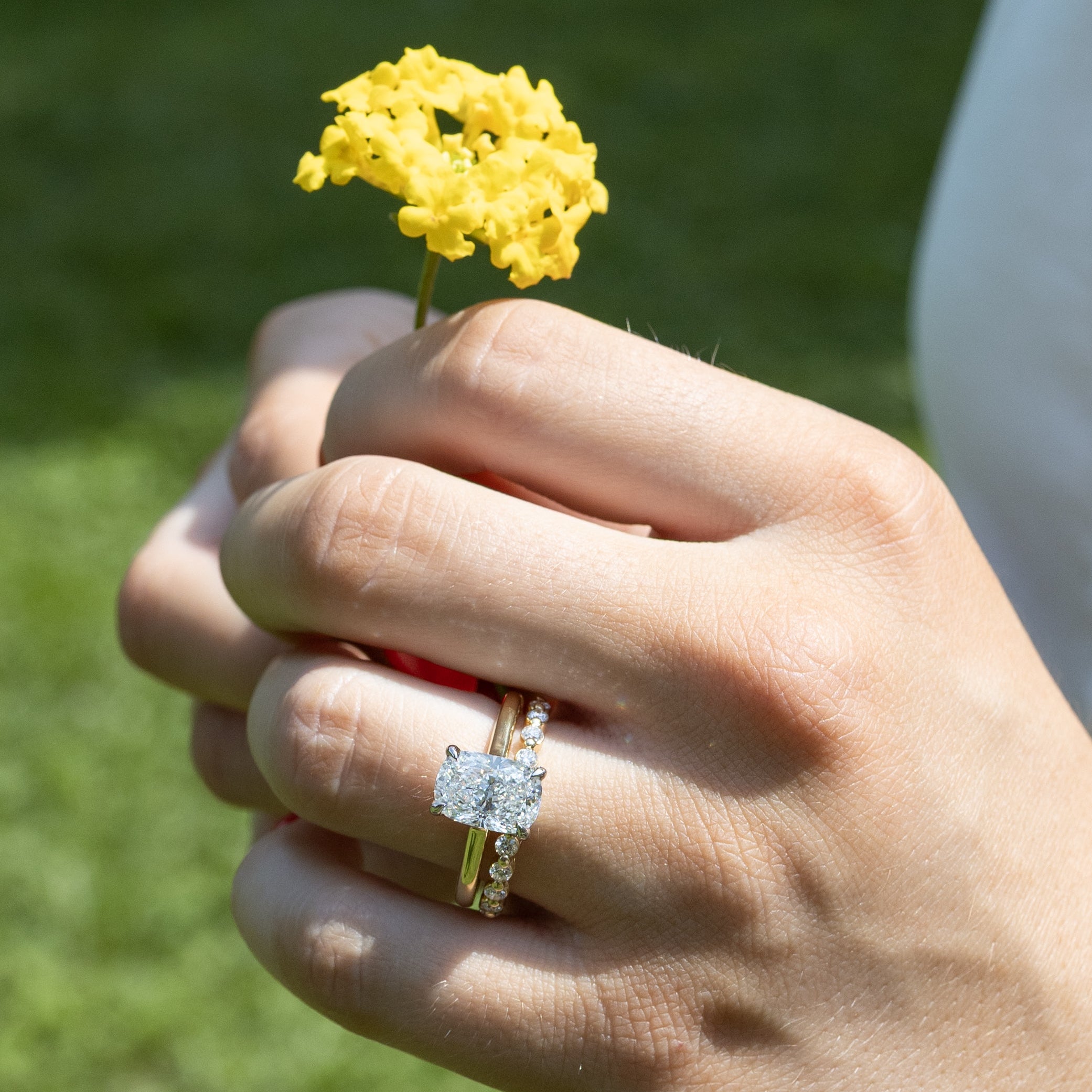 A close-up of a person's hand holding a small yellow flower outdoors, wearing a yellow gold ring with an elongated cushion-cut diamond and a matching yellow gold diamond band.