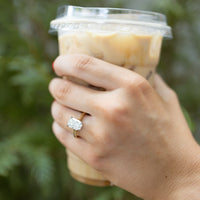 A close-up of a hand holding an iced coffee with a clear plastic cup lid, wearing a gold ring with a large elongated cushion diamond on the ring finger, against a blurred green background.