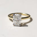 An elongated cushion cut diamond solitaire ring set in a yellow gold band, resting on a light marble surface with a soft shadow.