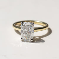 An elongated cushion cut diamond solitaire ring set in a yellow gold band, resting on a light marble surface with a soft shadow.