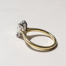 A solitaire engagement ring with a clear elongated cushion diamond in silver-toned setting on a polished yellow gold band, resting on a light marble surface.