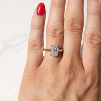 A close-up of a hand with red nail polish wearing a yellow gold ring with an elongated cushion diamond, set against a light marble background.
