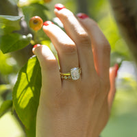 A close-up of a hand with red nail polish wearing a gold ring with a large elongated cushion diamond, set against a blurred outdoor background of green leaves and tree branches.