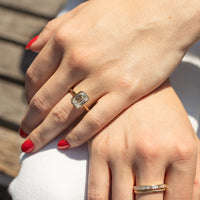 A close-up of a person's hands resting on white fabric, with a gold ring featuring a rectangular emerald-cut diamond on the upper hand. A second gold ring with small clear diamonds is worn on the lower hand.