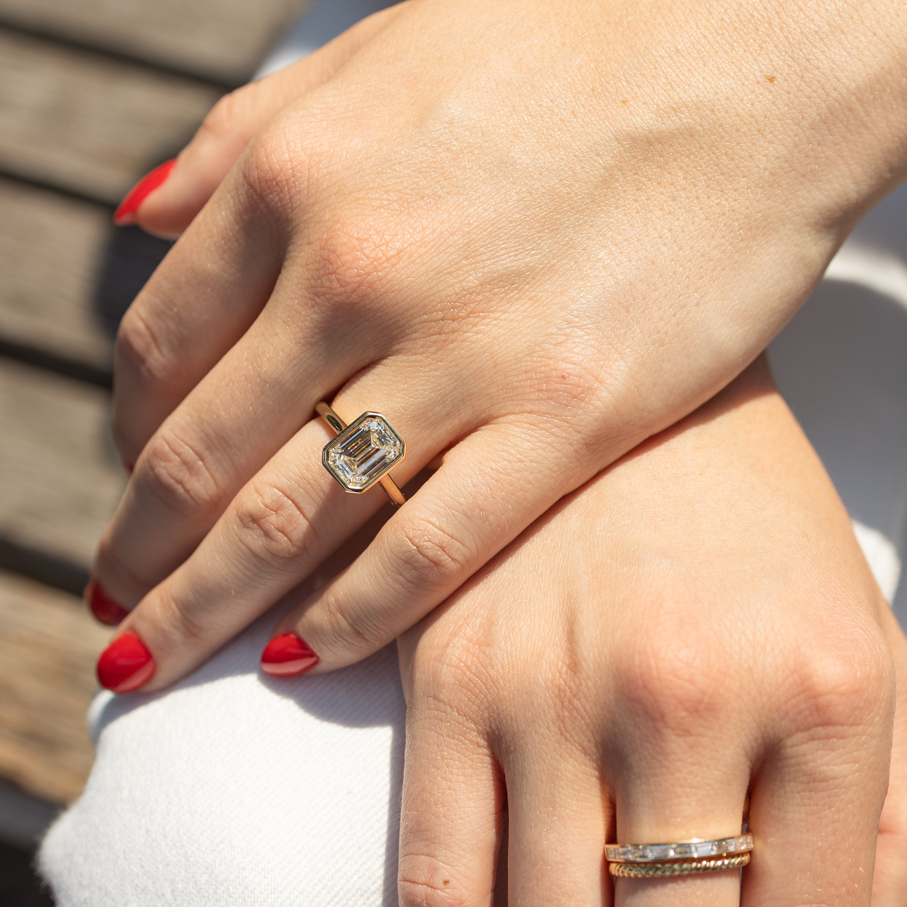 A close-up of a person's hands resting on white fabric, with a gold ring featuring a rectangular emerald-cut diamond on the upper hand. A second gold ring with small clear diamonds is worn on the lower hand.