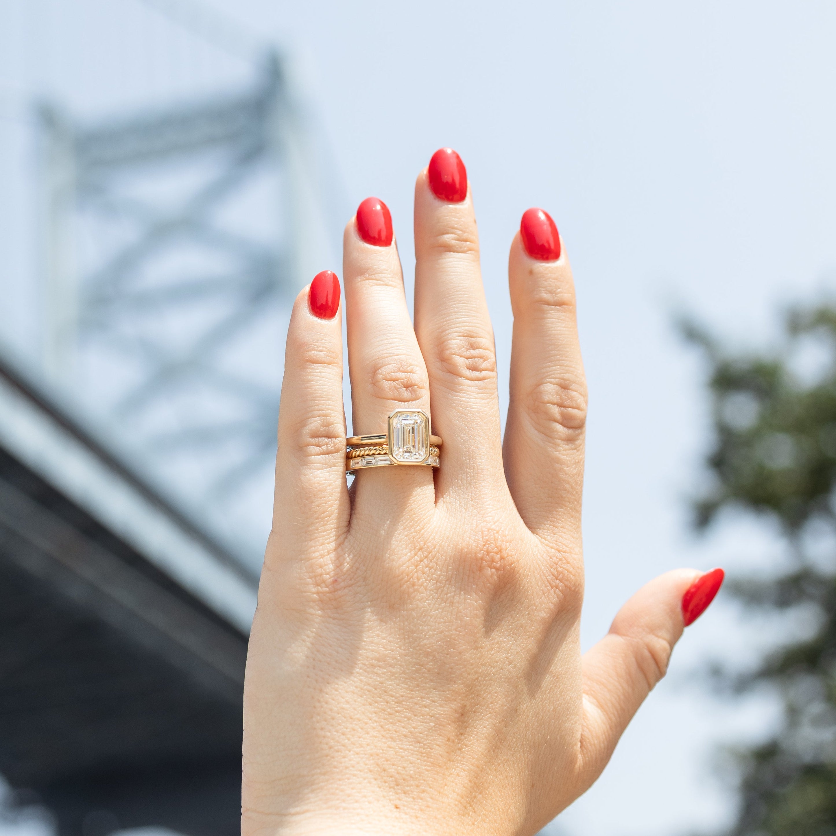A close-up of a person's hand with red nail polish wearing a gold bezel engagement ring with an emerald diamond, set against a blurred outdoor background of a bridge and sky.