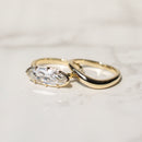 Two gold rings on a light marble surface, featuring a gold band with a moval-cut diamond and a second gold band with a smooth, polished curve.