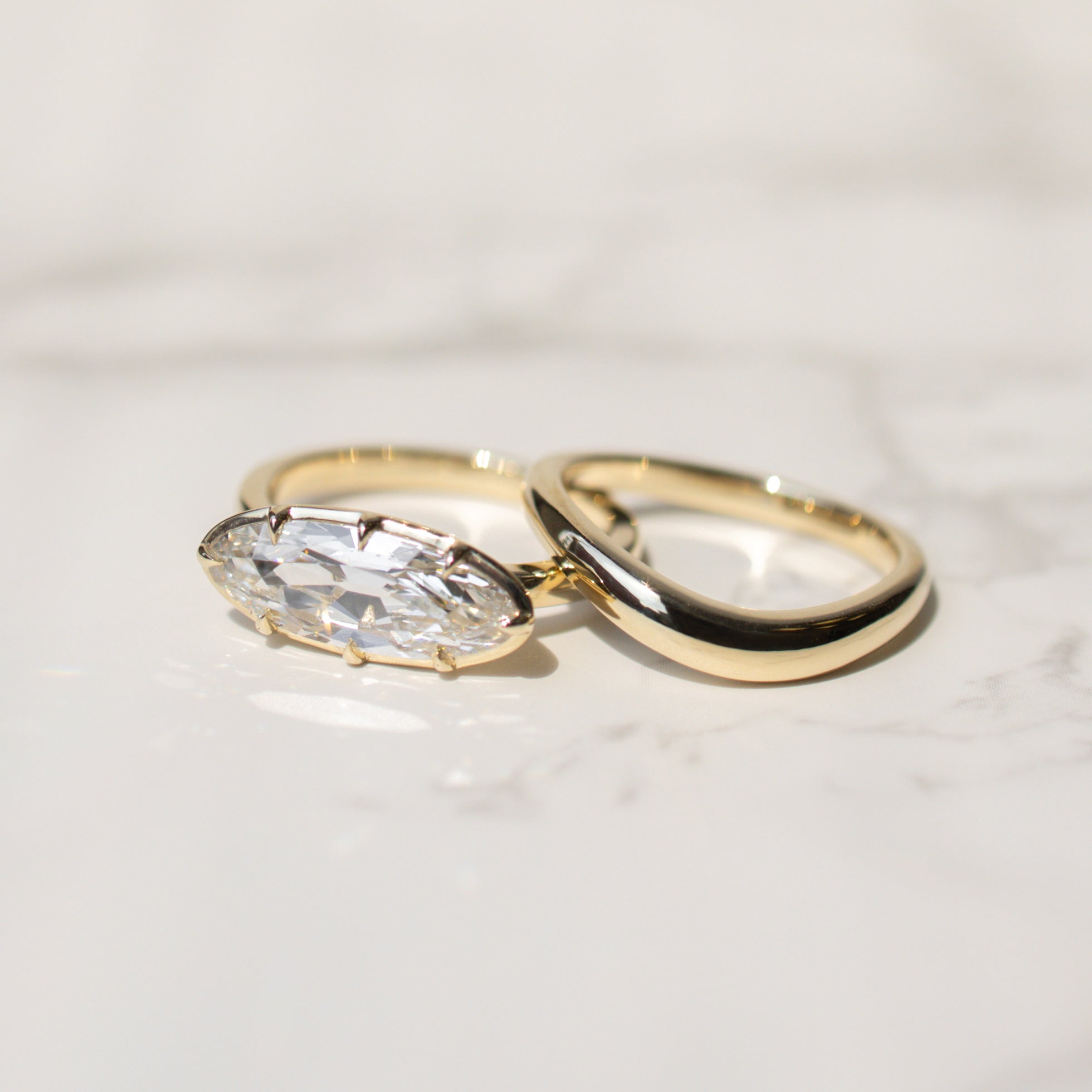 Two gold rings on a light marble surface, featuring a gold band with a moval-cut diamond and a second gold band with a smooth, polished curve.