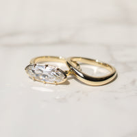Two gold rings on a light marble surface, featuring a gold band with a moval-cut diamond and a second gold band with a smooth, polished curve.