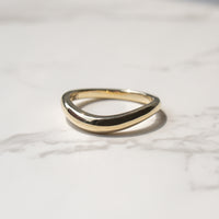 A polished gold wavy band ring resting on a light gray marble surface with a soft shadow.