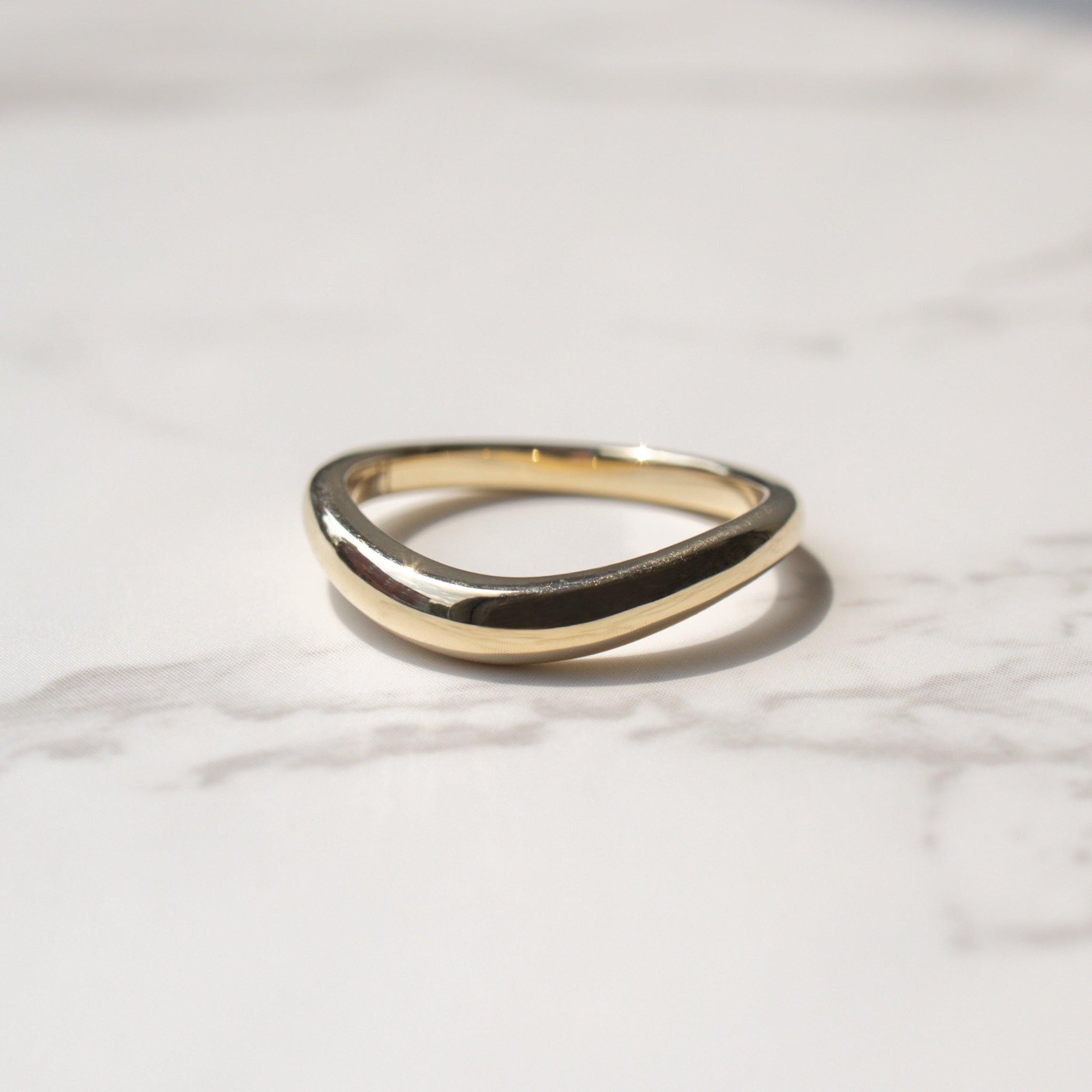 A polished gold wavy band ring resting on a light gray marble surface with a soft shadow.