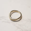A minimalist, polished curved gold band ring with a slightly tapered shape, resting on a light gray marble surface.