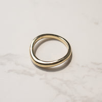 A minimalist, polished curved gold band ring with a slightly tapered shape, resting on a light gray marble surface.