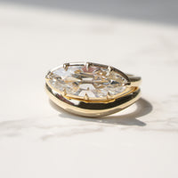 A gold ring featuring a large, georgian bezel-set moval-cut diamond, photographed on a light marble surface with soft shadows.
