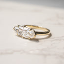 A gold ring with a moval-cut clear diamond in a georgian prong setting, resting on a light marble surface.
