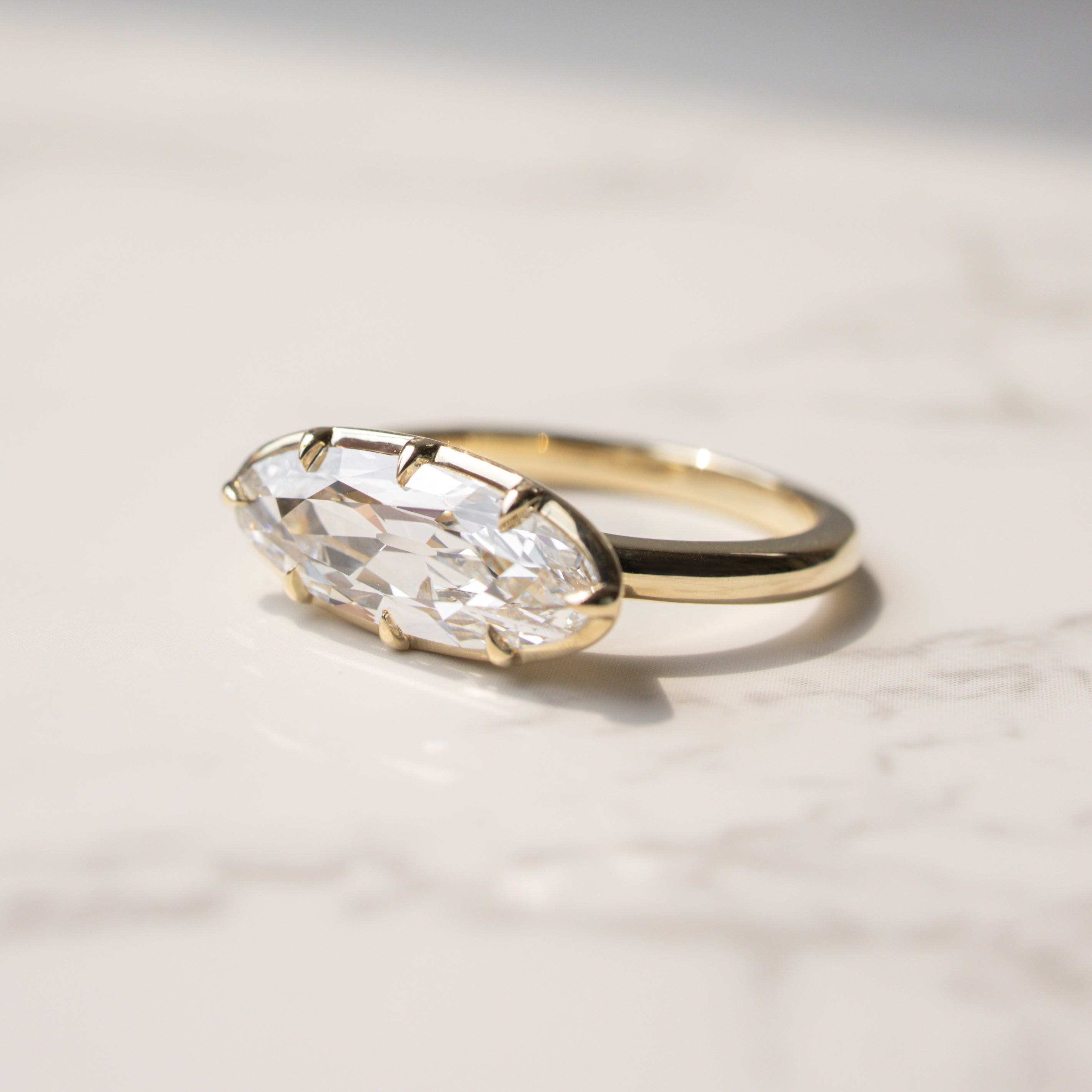 A gold ring with a moval-cut clear diamond in a georgian prong setting, resting on a light marble surface.