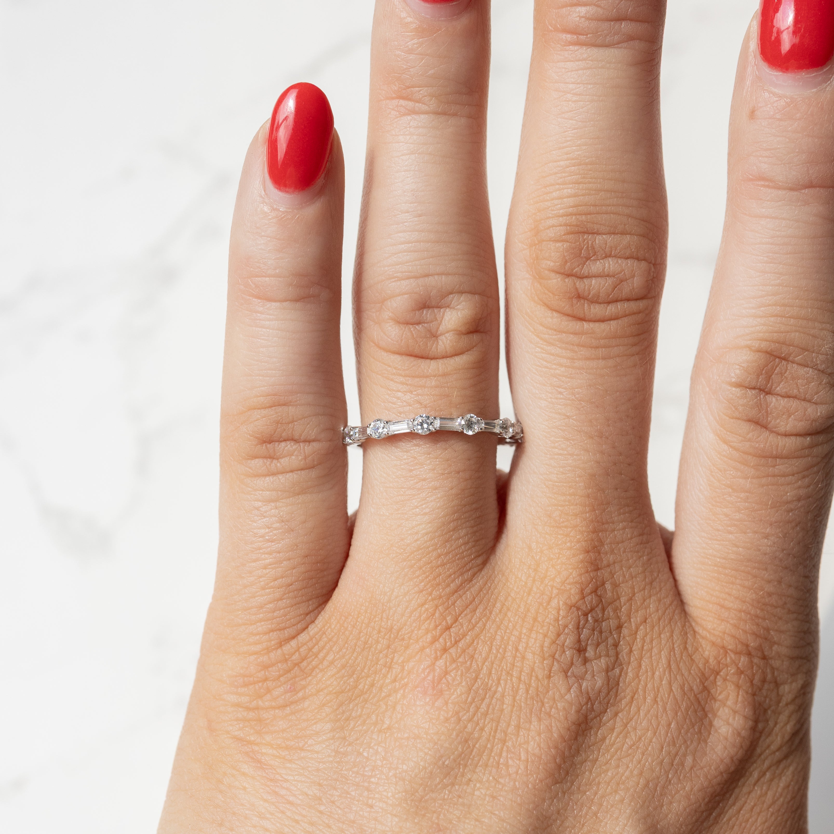 An on hand shot of a dainty white gold alternating baguette diamond wedding band. The hand has red fingernail polish.