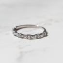 A dainty white gold diamond wedding band sitting on a white marble backdrop.   The band has alternating baguette and round diamonds.