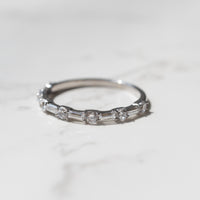 A dainty white gold diamond wedding band sitting on a white marble backdrop.   The band has alternating baguette and round diamonds.