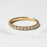 A yellow gold diamond wedding band sitting on a white marble backdrop. The band has small round diamonds going completely around it.