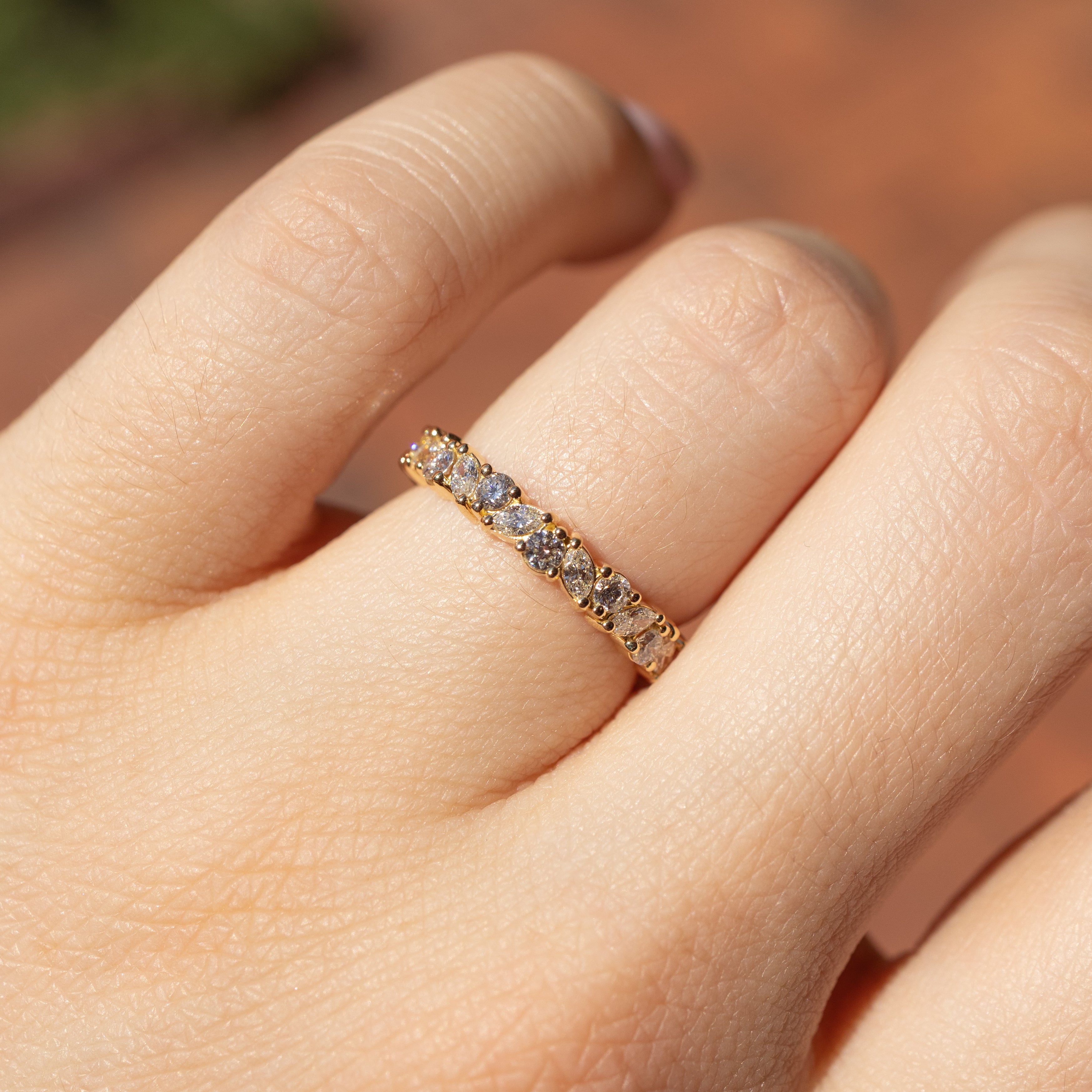 A close-up of a gold eternity ring with small clear stones worn on a finger. The ring features a continuous line of round-cut and marquise cut stones in a yellow gold setting, shown on a hand against a warm, blurred background.