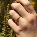 A close-up of a hand wearing a delicate gold band ring set with small round stones and marquise stones, photographed outdoors against a blurred natural background.
