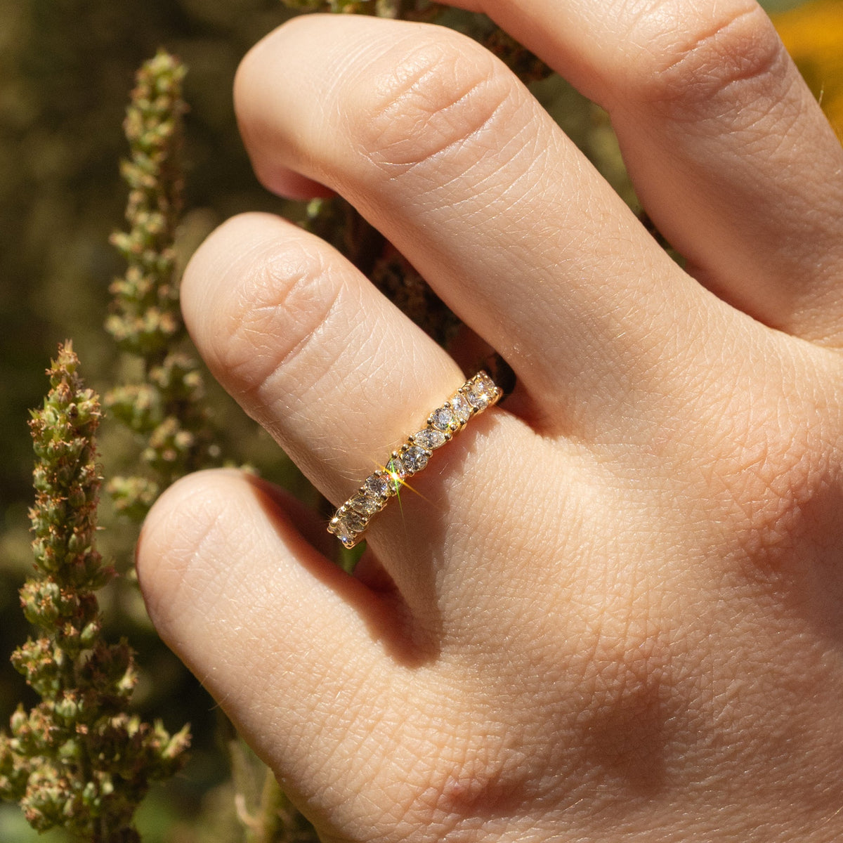 A close-up of a hand wearing a delicate gold band ring set with small round stones and marquise stones, photographed outdoors against a blurred natural background.