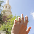 A close-up of a hand wearing a delicate gold band ring with small round and marquise diamonds, set against a blurred background of a church steeple and blue sky.