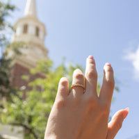 A close-up of a hand wearing a delicate gold band ring with small round and marquise diamonds, set against a blurred background of a church steeple and blue sky.