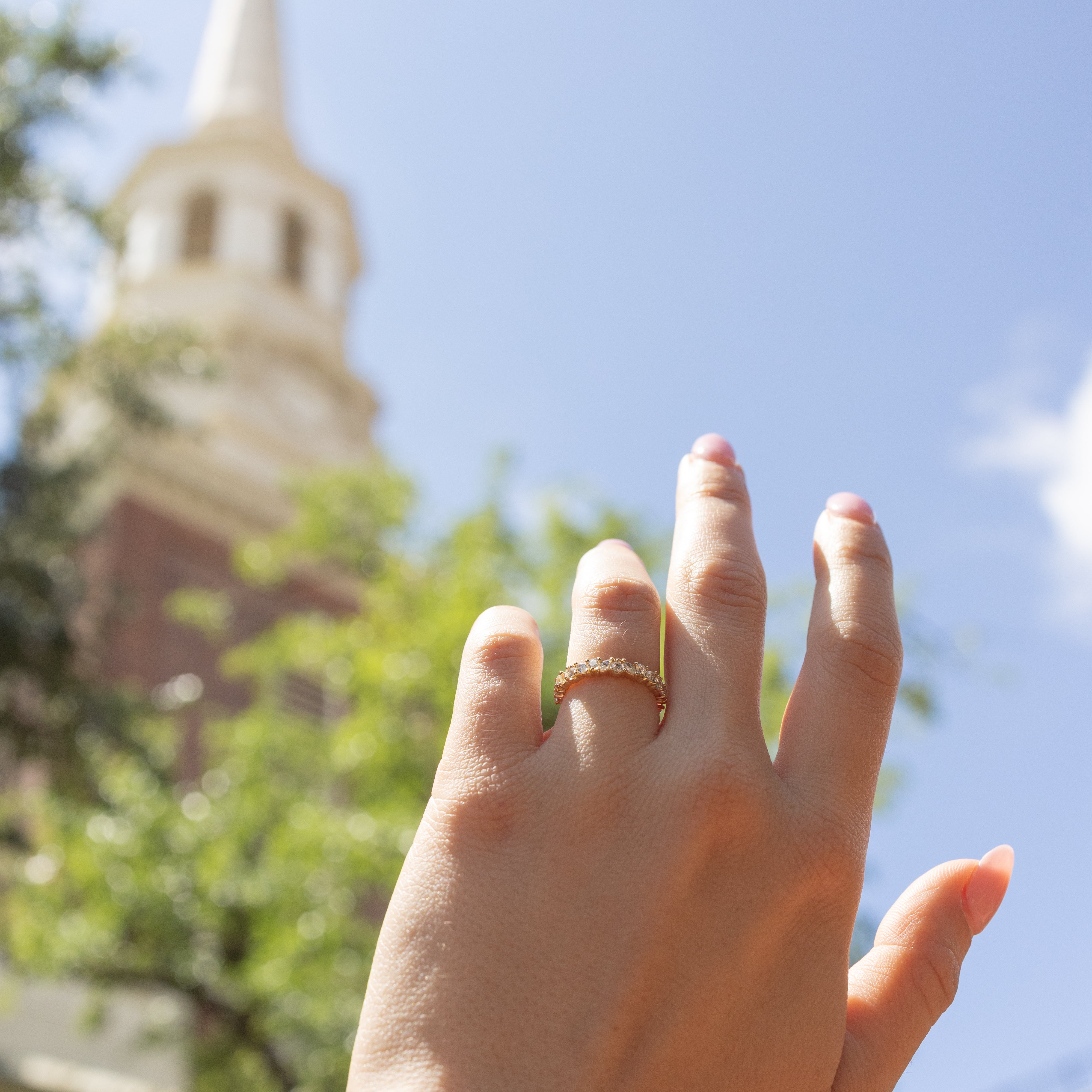 A close-up of a hand wearing a delicate gold band ring with small round and marquise diamonds, set against a blurred background of a church steeple and blue sky.