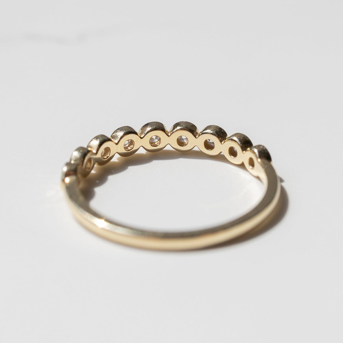 The back profile of a ring. It's a wedding band with small round bezel-set diamonds sitting on a white shadowy backdrop.