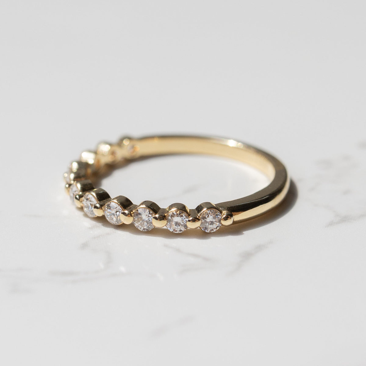 The side view of a yellow gold diamond wedding band with a row of small round diamonds sitting on a white marble backdrop.