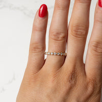 A close-up of a hand wearing a delicate gold eternity ring set with small round diamonds, shown against a light marble background.