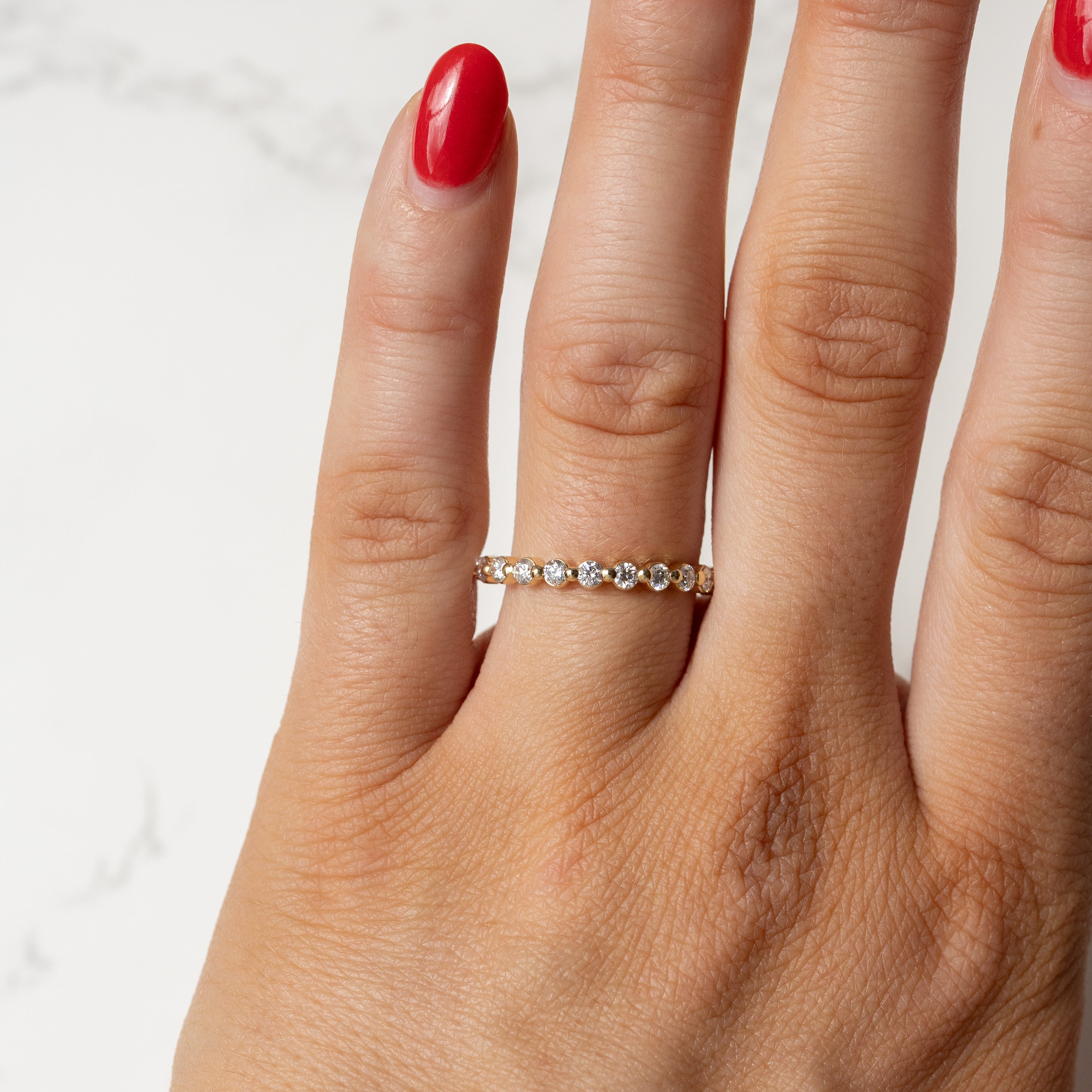 A close-up of a hand wearing a delicate gold eternity ring set with small round diamonds, shown against a light marble background.