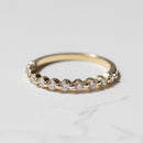 A yellow gold wedding band with a row of small round diamonds with a shared prong setting sitting on a white marble backdrop.