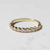 A yellow gold wedding band with a row of small round diamonds with a shared prong setting sitting on a white marble backdrop.